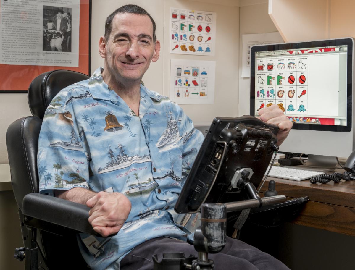 George is wearing a printed blue shirt. He has an augmentative and alternative communication (AAC) device mounted to his wheelchair and there is a computer on the desk displaying communication software.