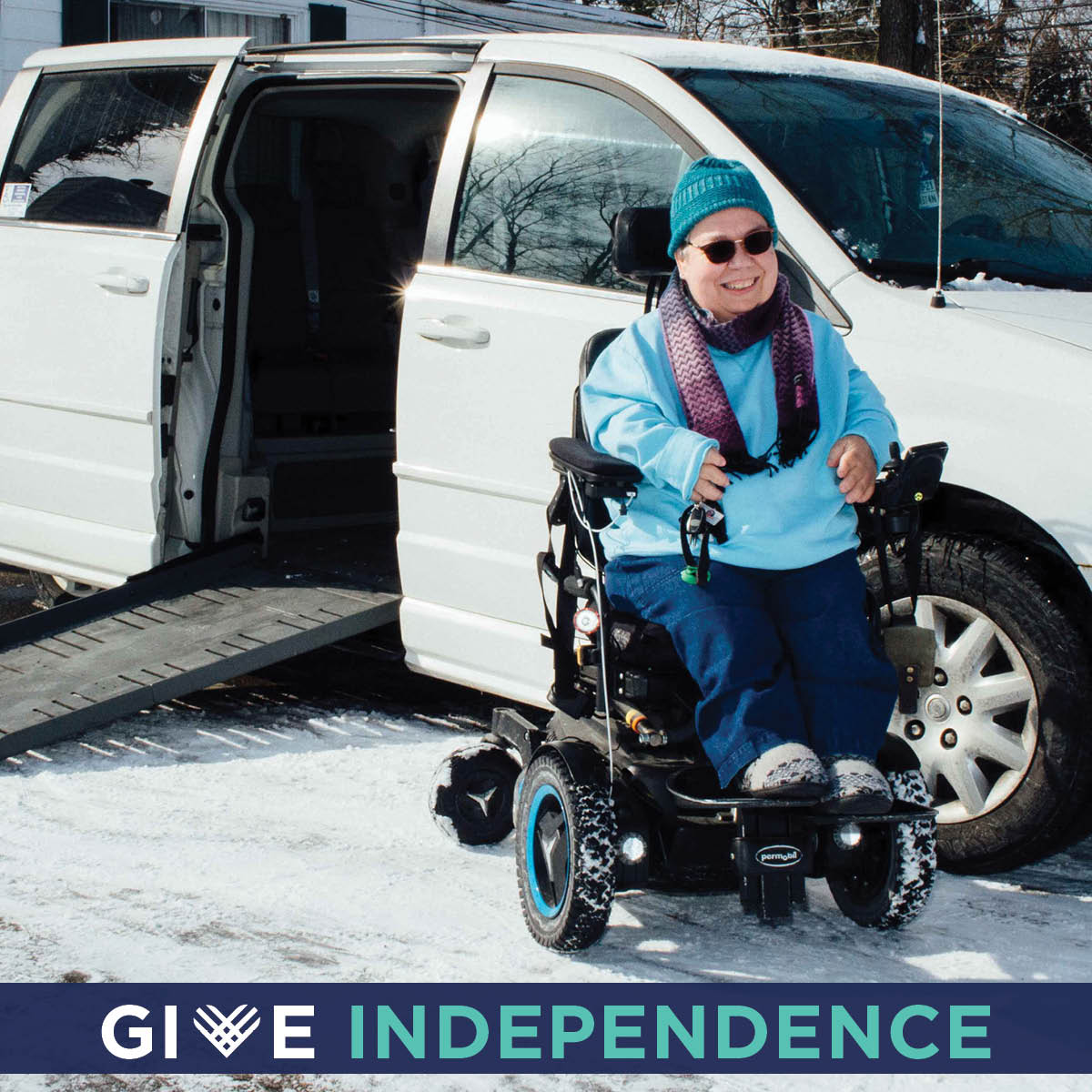Woman sits smiling in her power wheelchair in front of her adapted van with the ramp outstretched. Give independence is written across the bottom.
