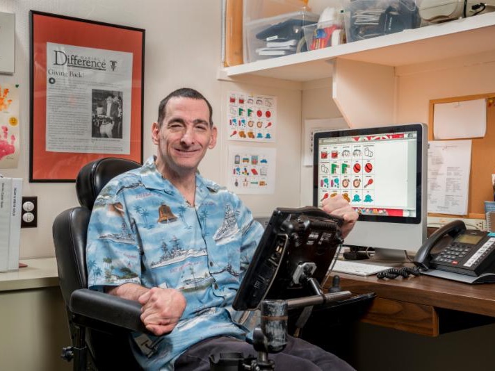 George is a wheelchair user, wearing an aqua blue button up shirt, at a desk, with framed pictures on the wall