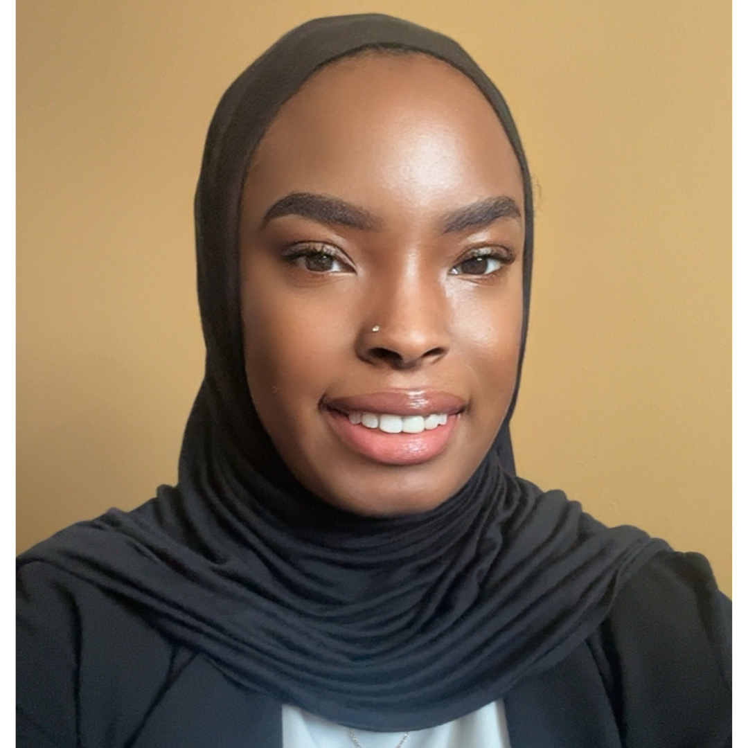 A black woman wears a black head covering, gray shirt, and black jacket. 