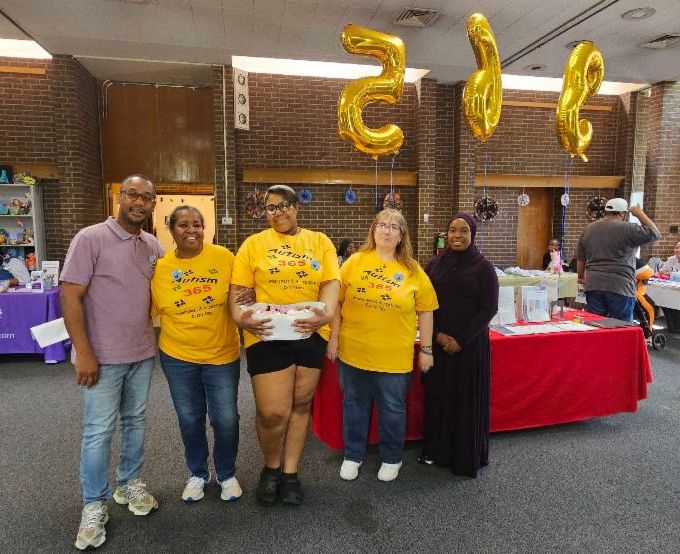 There are five people standing in front of "365" balloons. Quintin is standing on the left, Laquesha is standing on the right, and there are three women wearing yellow shirts standing in the middle.