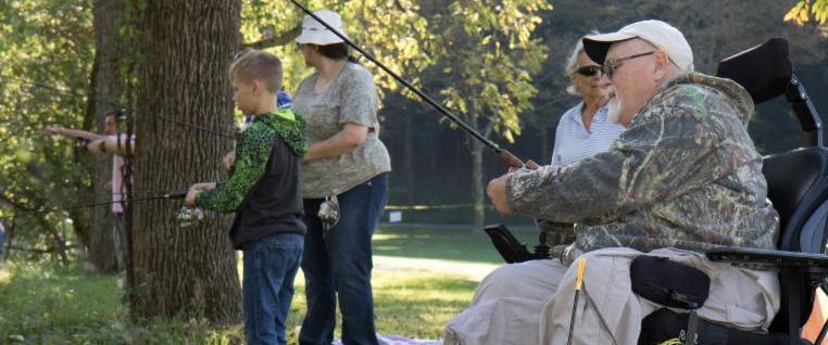Older man sits in a his power wheelchair adjusting his fishing rod. Two women and a boy are behind him in the grass_ the boy is also fishing.