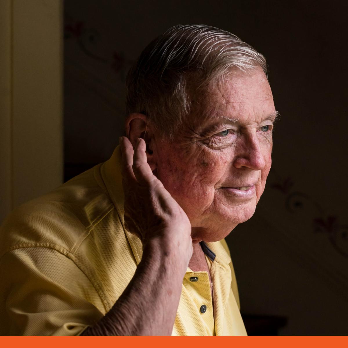 An older man gently holds his hand to his ear.