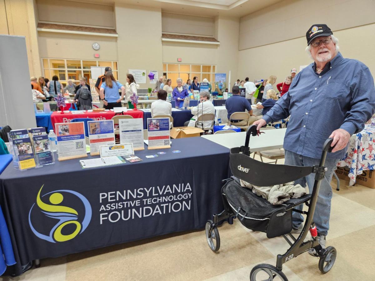 There is a man wearing a blue shirt, jeans, and a black hat. He is using a walker and standing in front of PATF's resource table.