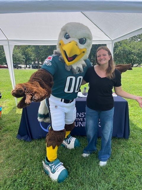 Swoop is the Philadelphia Eagles' mascot and Mary Kate is a woman wearing a black shirt. They are standing in front of PATF's table.