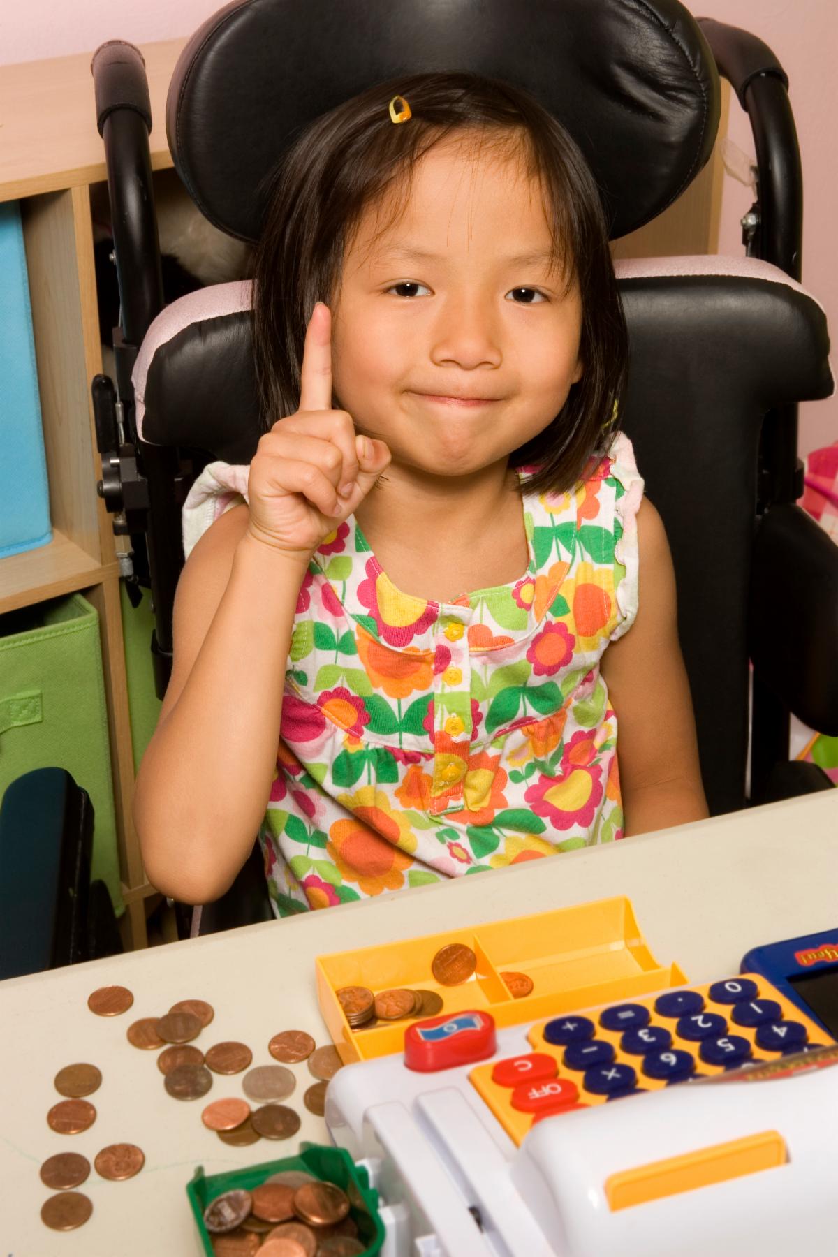 A young girl sits at a table counting coins near a toy cash register.