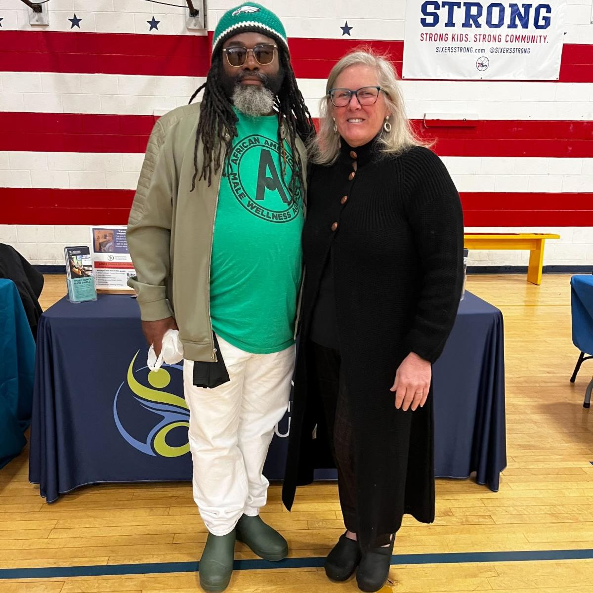 There is a man on the left wearing a green shirt with a green jacket, white pants, and a green Eagles hat. On the right, Wendy Davis is wearing a black shirt, black jacket, and black pants. They are standing in front of PATF's resource table.