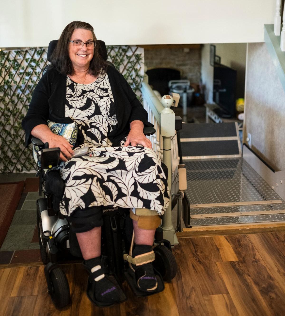 Colleen is a white woman and is wearing a black and white dress. She is sitting in her wheelchair next to the inclined platform lift.