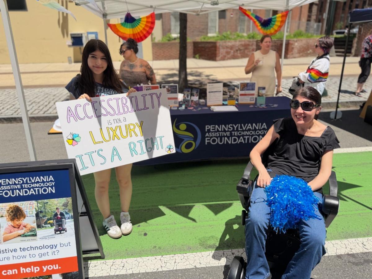 There is a young girl standing in front of PATF's resource table holding a sign that says, "Accessibility is not a luxury, it's a right." Next to her, there is a woman sitting in her wheelchair holding a blue pompom.