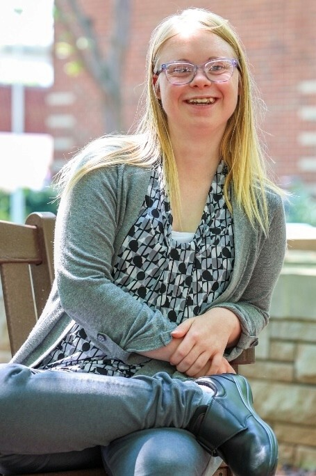Sara has blonde hair and glasses. She is smiling and wearing a shirt with a design on it and a gray cardigan.