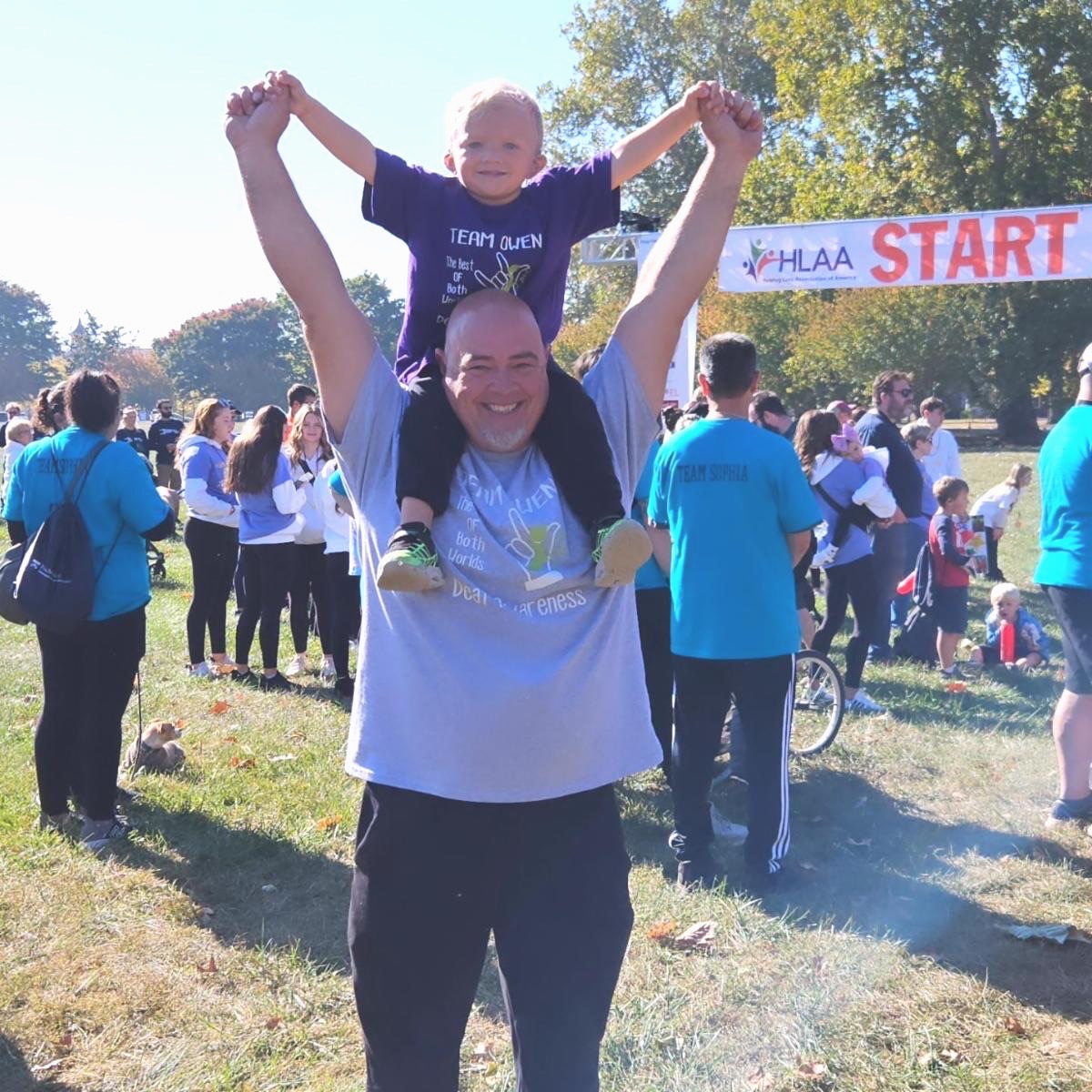 A man has a young boy seated on his shoulders and their hands are raised.