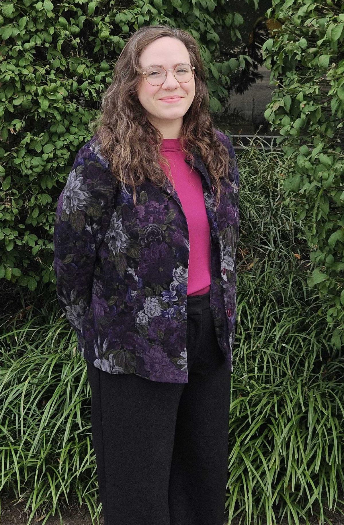 Rachelle has long brown curly hair. She is wearing a pink shirt with a flower printed black cardigan over top and black pants. She is wearing glasses. She is standing in front of bushes outside.