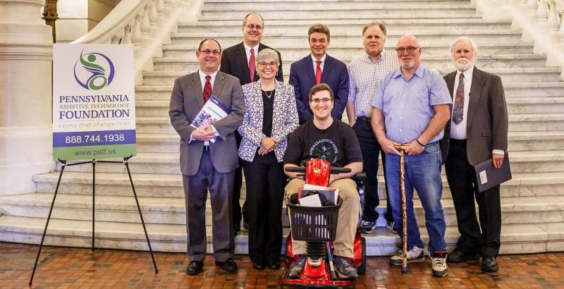 8 people pose next to a PATF poster in front of a marble staircase_ one of them seated in a red scooter_ the rest standing.