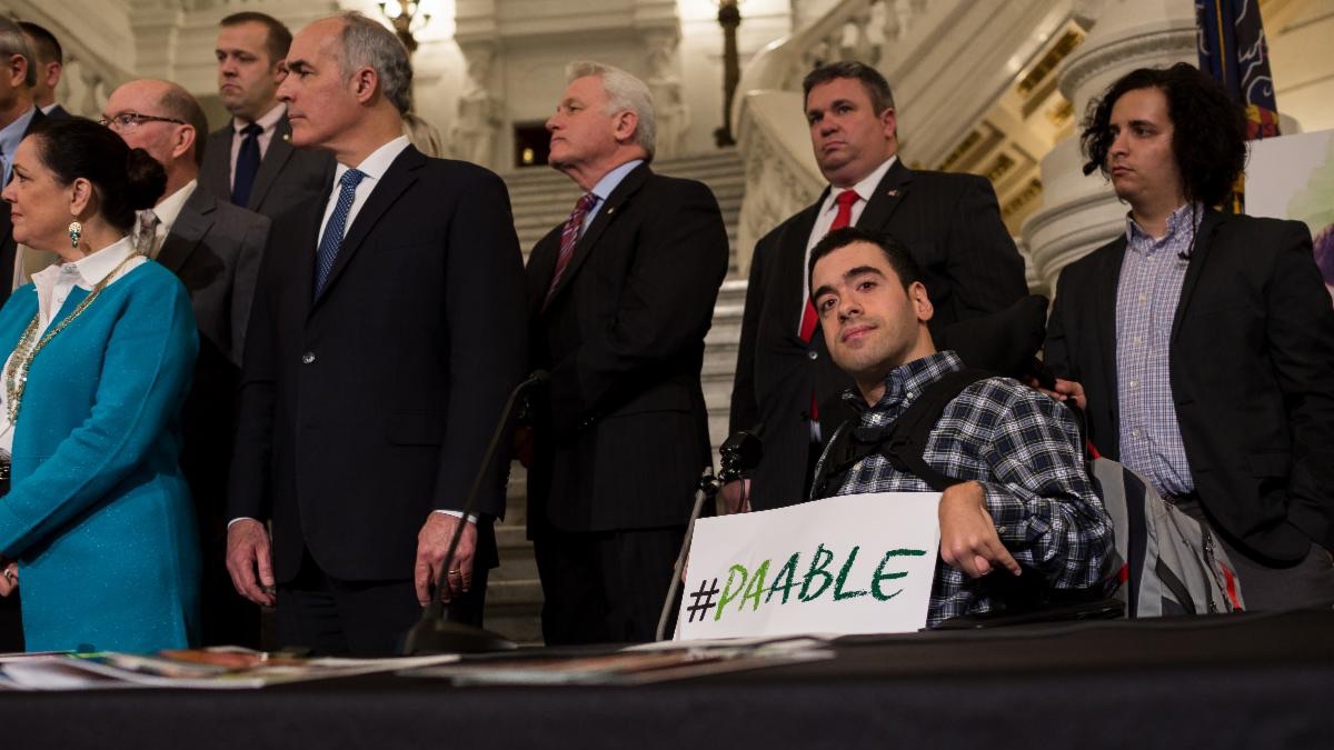 A white man in a wheelchair is in front of several people while holding a PA ABLE sign.