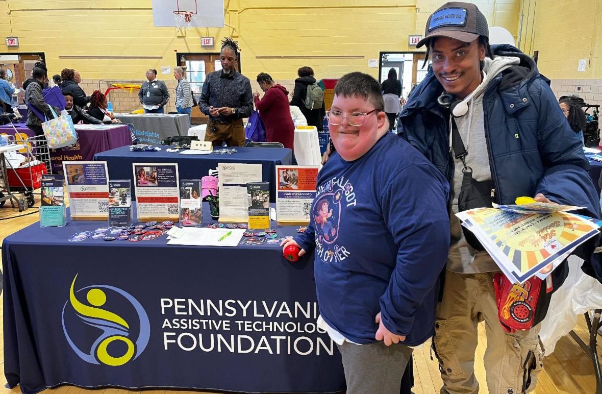 There are two men smiling and standing together in front of PATF's resource table in a crowded gymnasium.