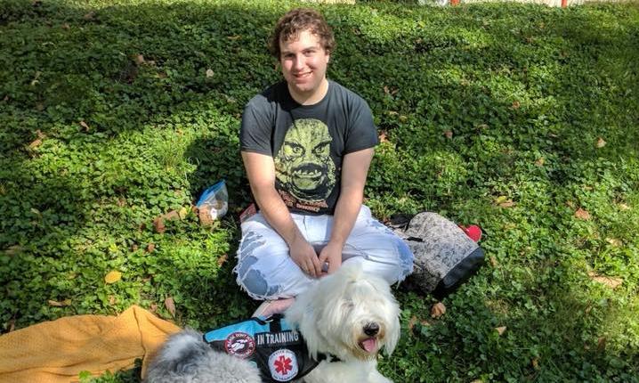 Young man sits in the grass with his service dog_ a sheepdog wearing a training vest.