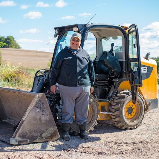 A man smiles and stands by a side entry skid steer with rolling farmland behind him.
