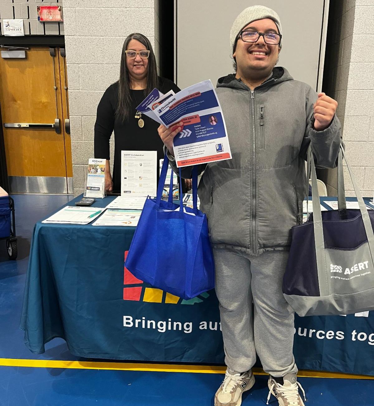There is a man wearing a gray jacket, jeans, and a tan hat. He is holding two bags and several resource papers and standing in front of a resource table. There is a woman standing behind the table.