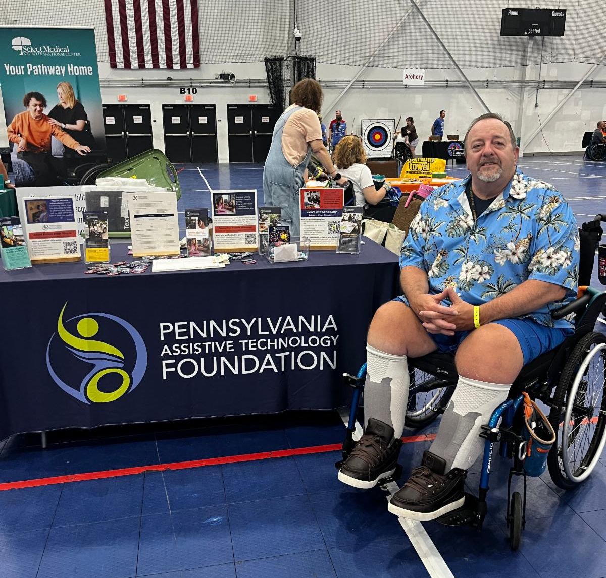There is a man sitting in his wheelchair in front of PATF's table. He is wearing a blue floral shirt and blue pants.