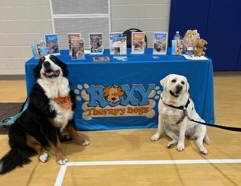 There are two therapy dogs sitting on the side of the "Roxy Therapy Dogs" resource table.