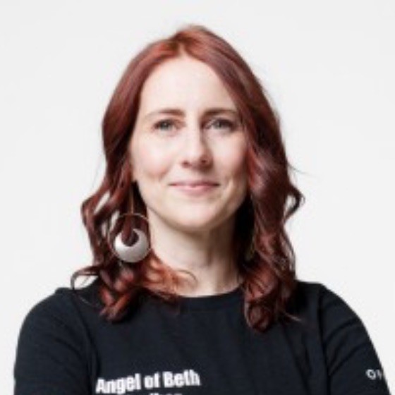 Bethany is a white woman with curled red hair. She is wearing a black shirt.