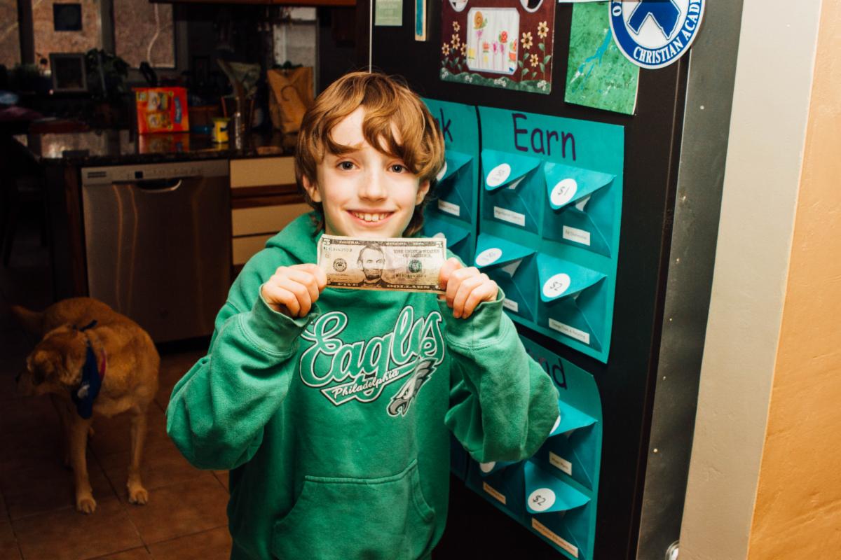 A young boy wearing a green sweatshirt smiles while holding a five dollar bill.