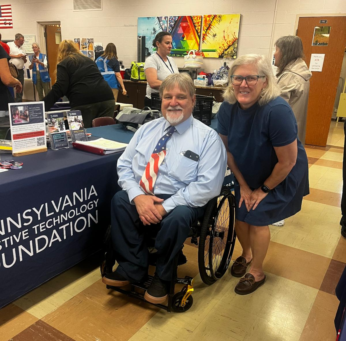 Wendy Davis is wearing a blue dress. She is standing next to a man who is sitting in his wheelchair. He is wearing a blue button up shirt with an American flag tie and dark blue pants. They are in front of PATF's resource table.