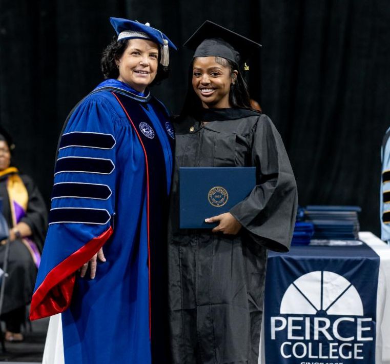 Shideerah is wearing a black gown and cap and standing on stage holding her diploma. She is standing next to a woman wearing a blue gown and cap. There is a table that says, "Peirce College" next to Shideerah.