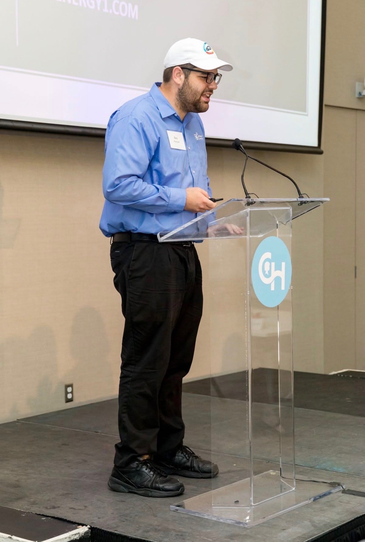Ben is standing at a podium. He is wearing a blue collared shirt, black pants, and a white baseball cap.