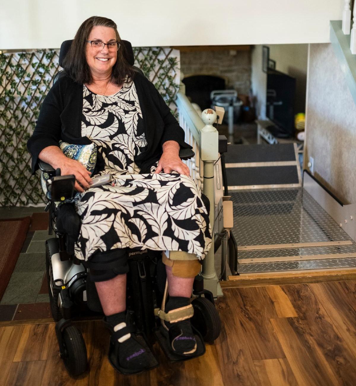 A white woman wearing glasses and, a black and white dress, is in a power wheelchair next to an inclined platform lift.