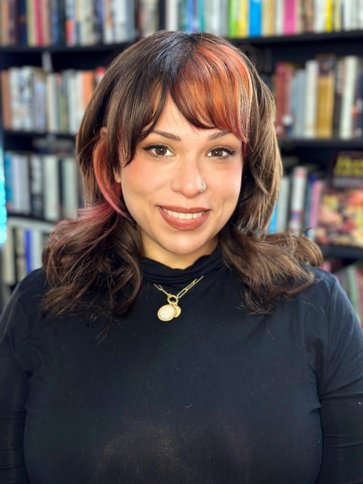 Wendy has shoulder length brown hair with orange highlights. She is wearing a black shirt and gold necklace. There is a bookshelf behind her.