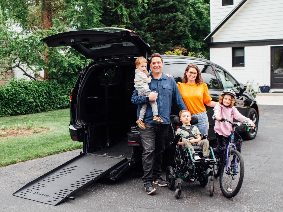 There is a man in a jean shirt standing up holding his young child. A woman in an orange shirt is standing next to him. There is a young girl sitting on her bike and wearing a helmet. There is a young boy sitting in his wheelchair. They are all standing in front of their adapted vehicle.