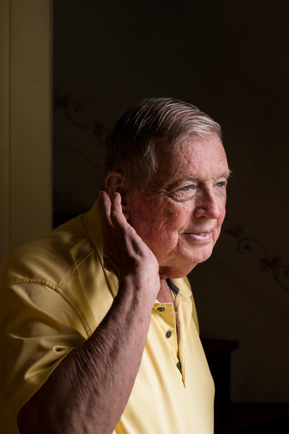 Bob is an older white man. He is wearing a yellow shirt and is holding his hand up to the hearing aid in his ear.