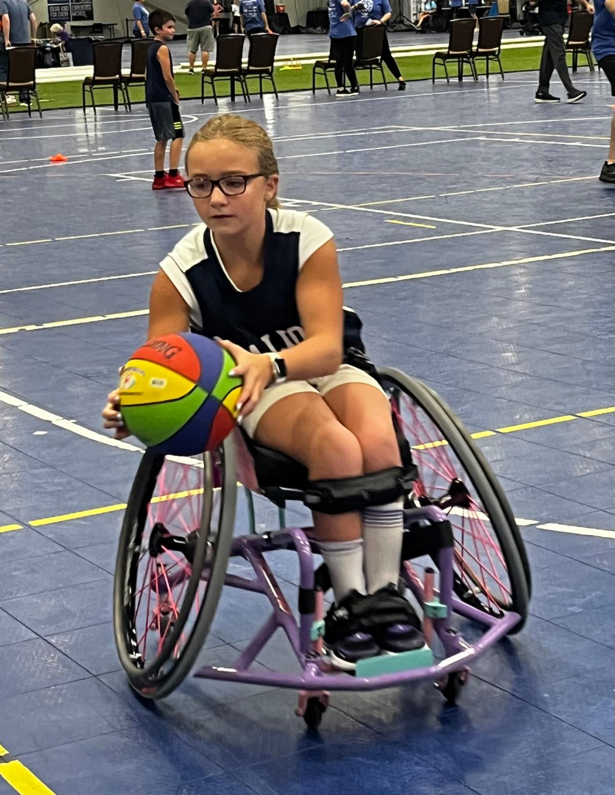 There is a young girl wearing glasses and is sitting in her wheelchair. She is holding a colorful ball.