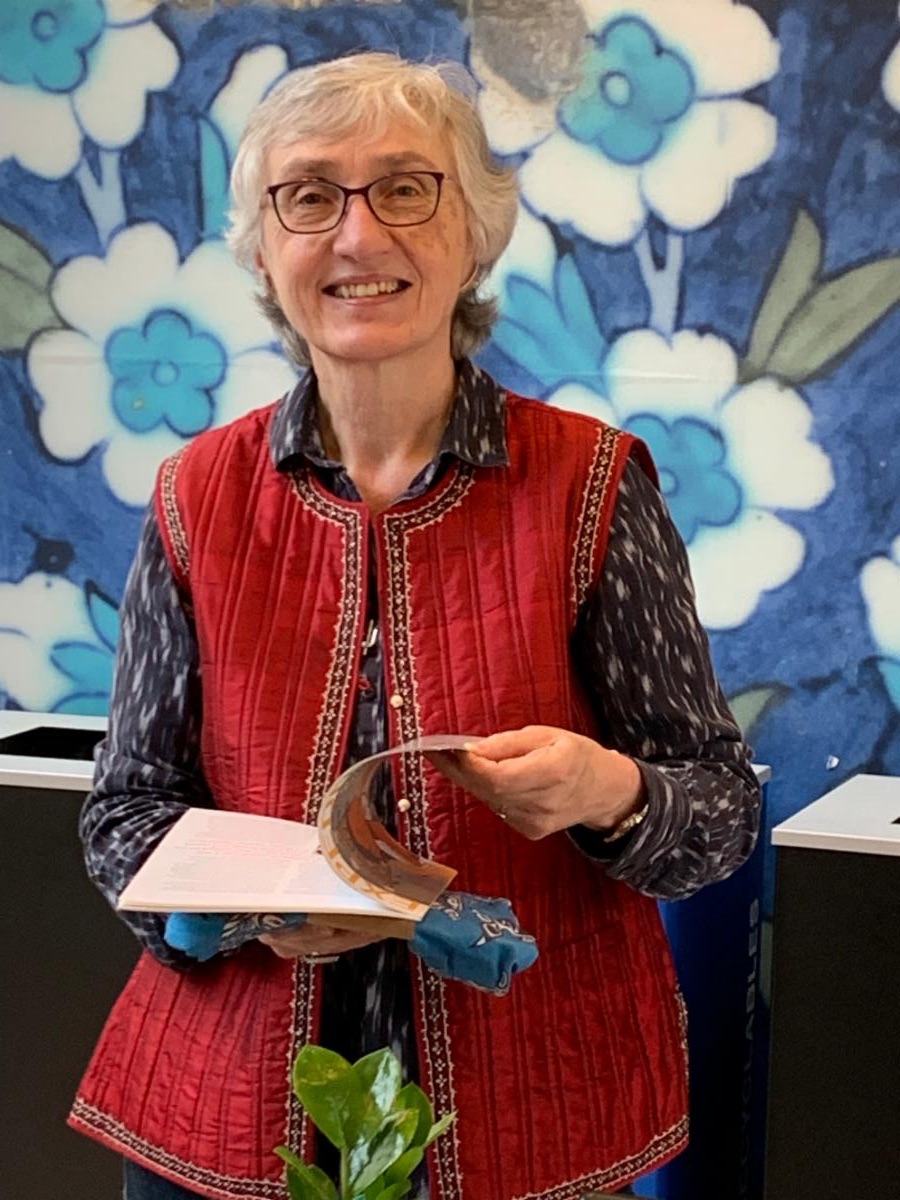 Marlies Singh is an older woman with short grayish hair, wearing glasses, a buttoned down shirt, and a red vest. She is smiling and holding an opened book.