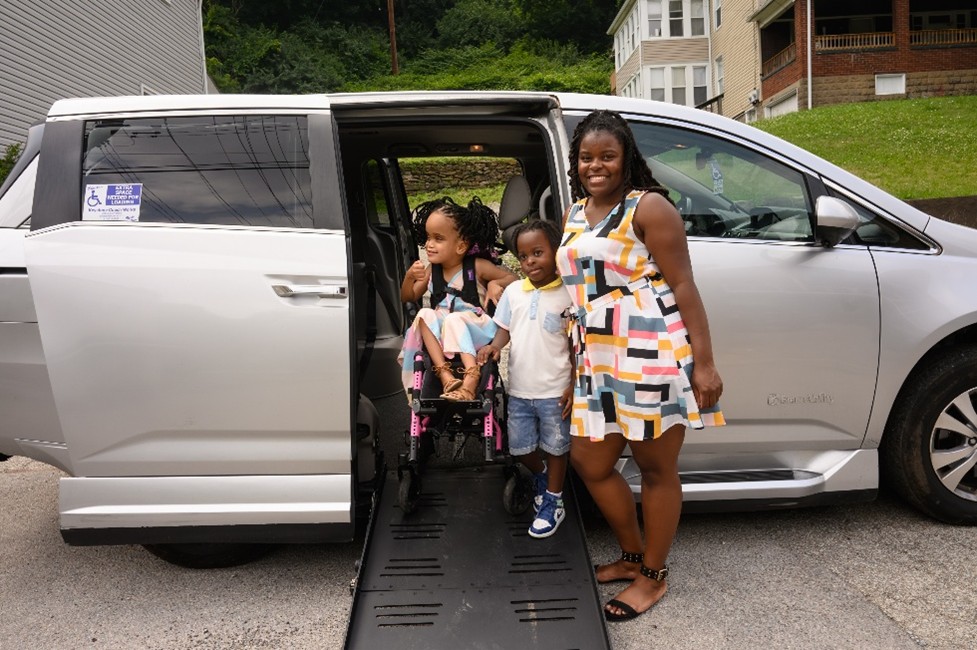 A smiling woman is standing next to a silver accessible minivan with its side ramp deployed. A young girl in a wheelchair and a young boy are on the ramp.