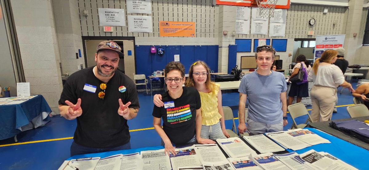 A man with a hat on, a woman with short brown hair, a girl with long light brown hair, and a man with brown hair standing behind a table smiling.