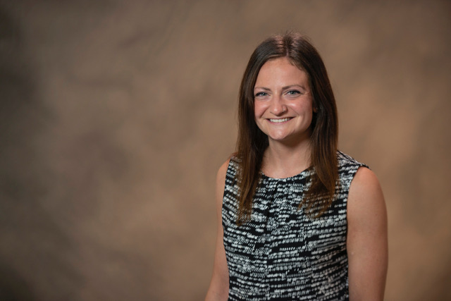 Mary Kate, a young white woman, has long brown hair, smiling, wearing a sleeveless black and white shirt, standing in front of a brown background.