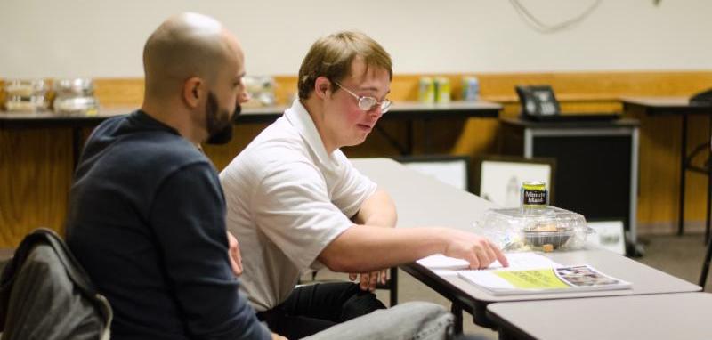 Two men sit at a table_ one man points to a workbook on the table in front of him.