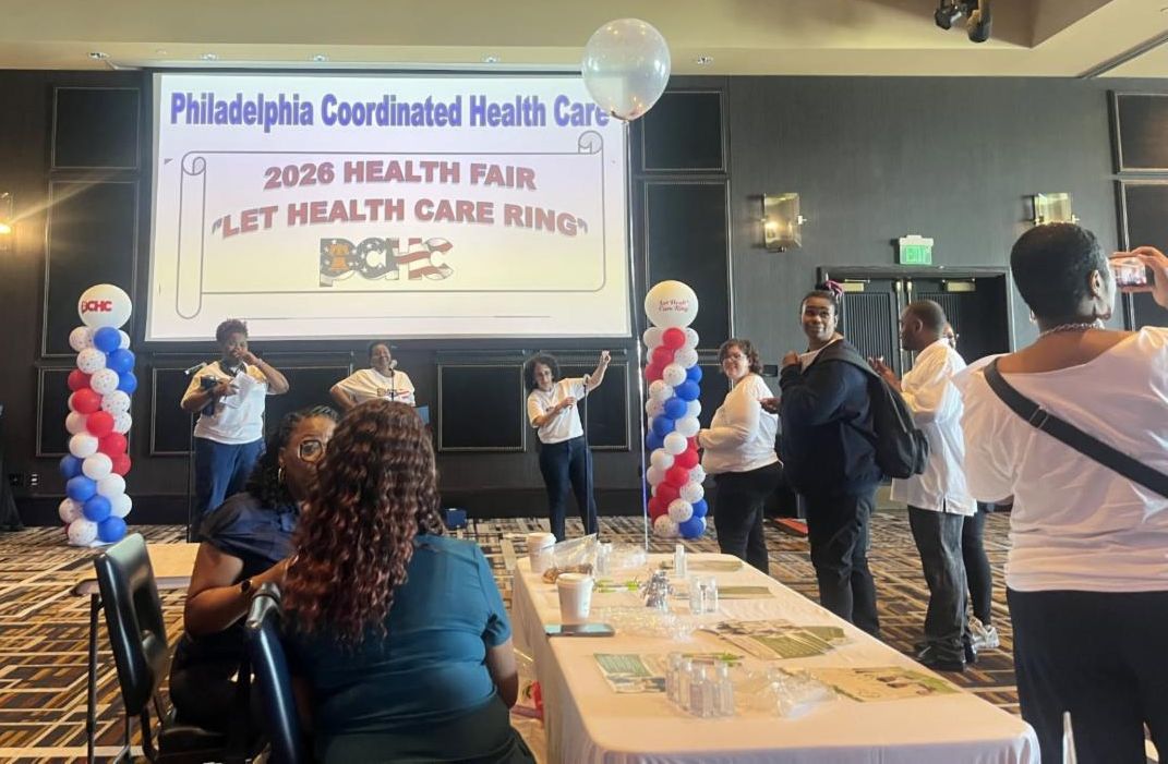 There is a projector that says, "Philadelphia Coordinated Health Care 2026 Health Fair 'Let Health Care Ring' PCHC." There are red, white, and blue balloon towers next to it. Attendees are standing around.