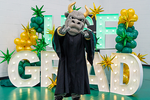 Rocky D. Bull holding the horns up sign with his graduation gown and balloons behind him 