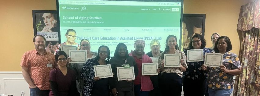 A group of palliative care trainers and assisted living staff stand together in a classroom holding certificates of completion. A presentation slide for the Palliative Care Education in Assisted Living Lab is displayed on a screen behind them