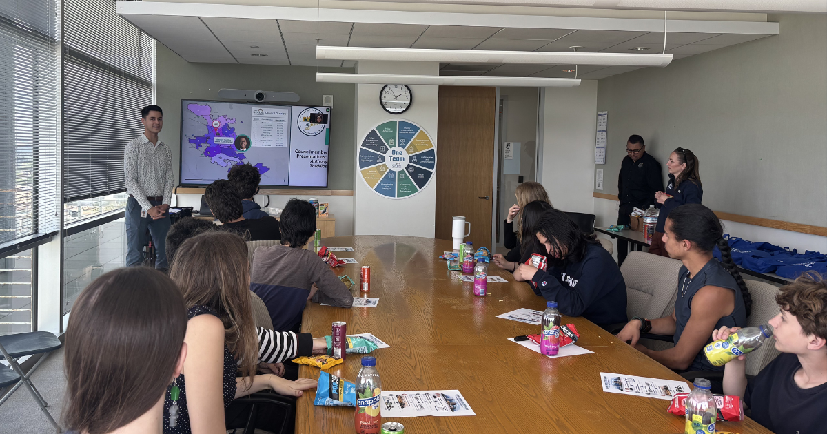 Councilmember Anthony Tordillos addressing approximately a dozen Hillbrook High students gathered around a conference table, with a map of San José District 3 displayed on a screen behind him.