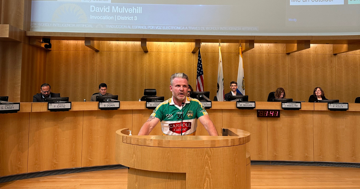 David Mulvehill wearing an Irish rugby jersey, standing at a podium, addressing the City Council Chambers