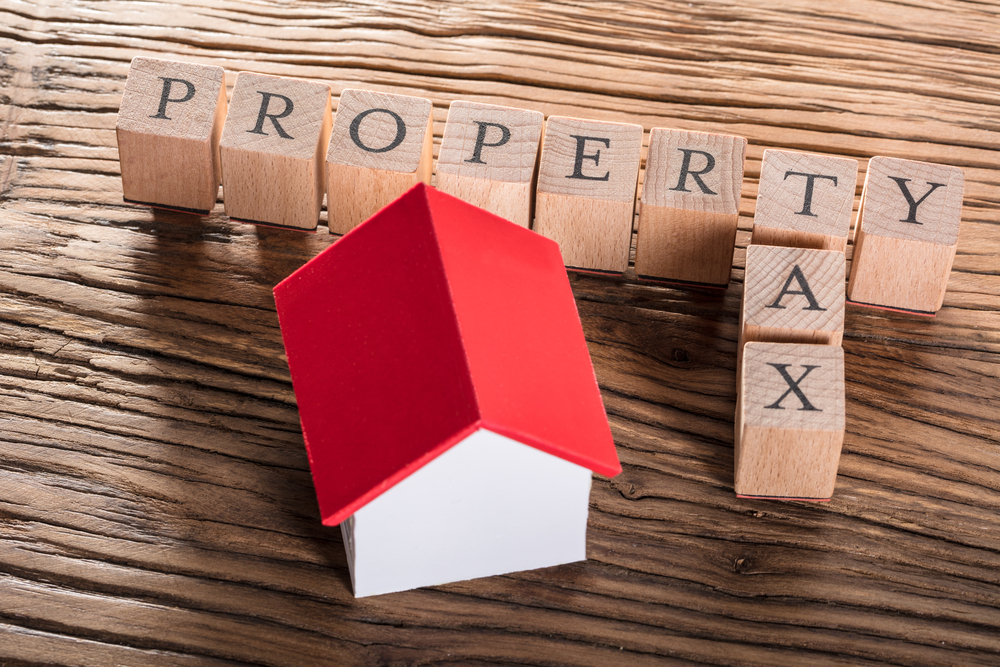 White toy house with red roof placed on wooden floor with words property tax written above it