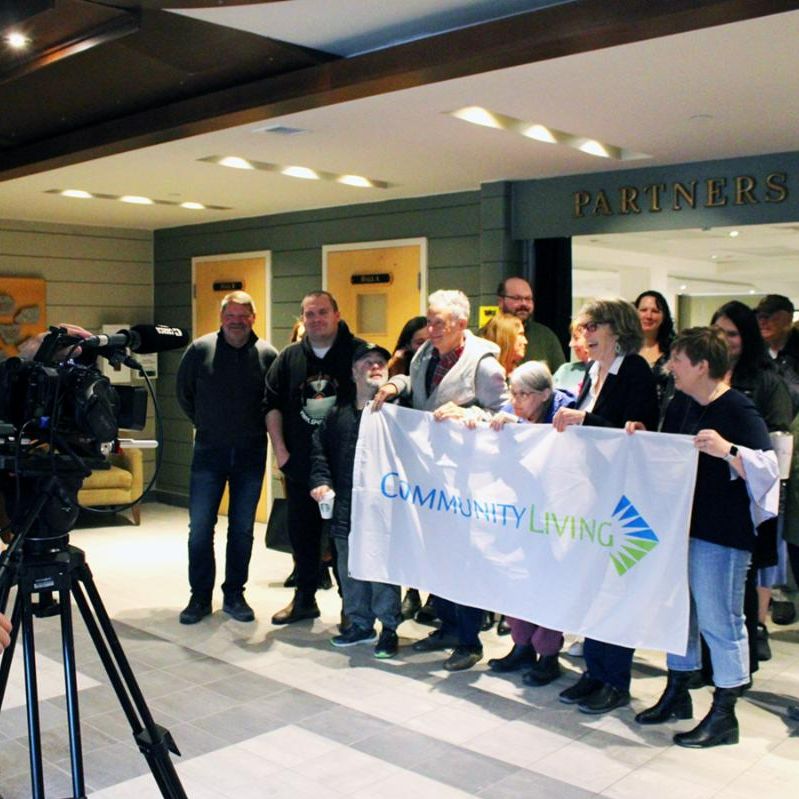 A large group of people holding a six-foot-by-three-foot flag of the Community Living logo on a white background.