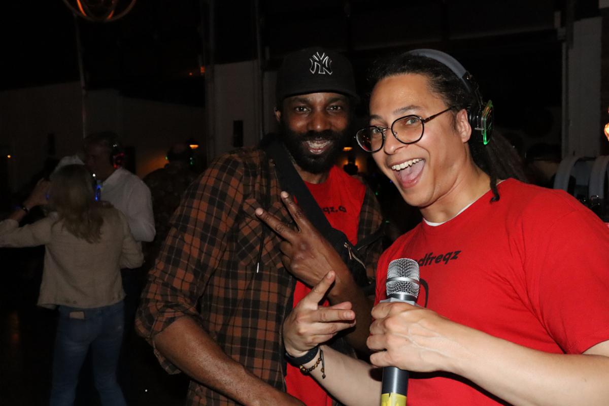 2 men give peace signs while holding a microphone and wearing wireless headphones