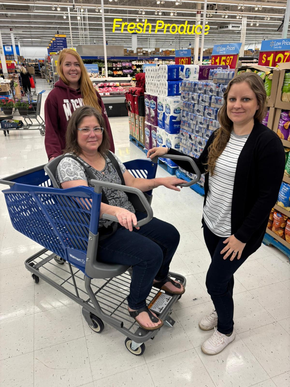 3 women in a grocery store with 2 standing around a modified shopping cart and 1 seated in the full sized seat built into it