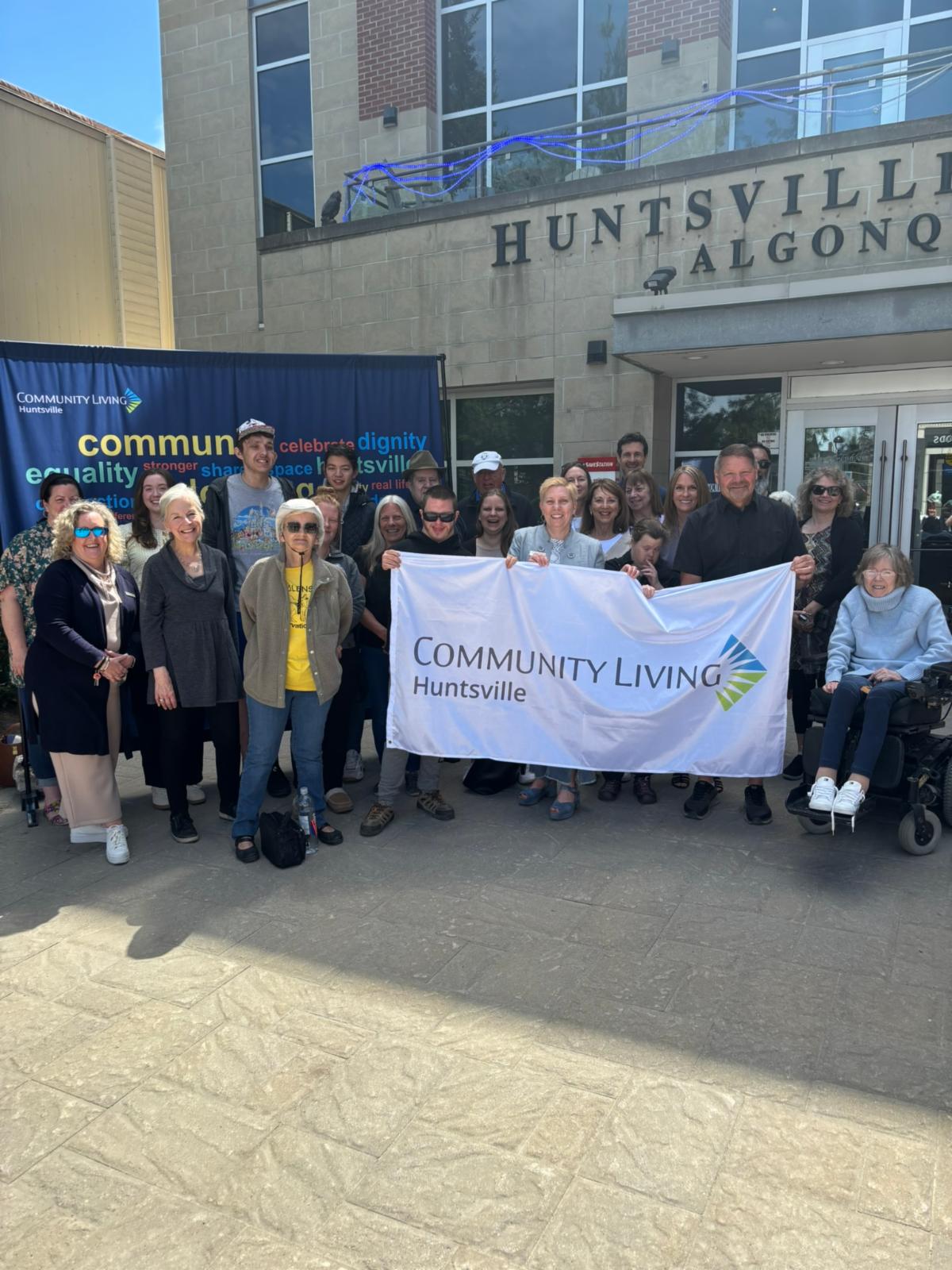 A group of people hold a white Community Living flag while standing in front of Huntsville Civic Centre
