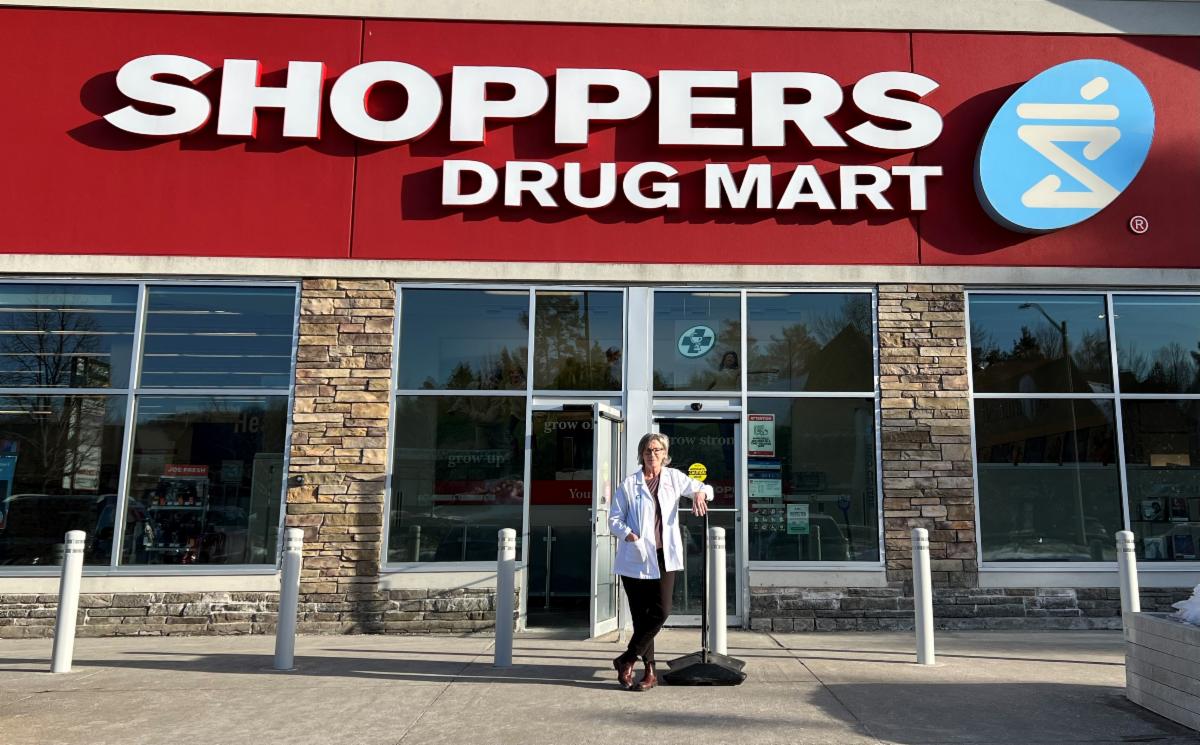 A woman standing in front of a building that has a large red and white sign over the front door that reads Shoppers Drug Mart